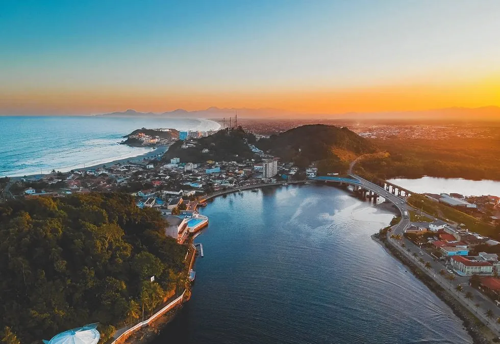 Vista aérea de uma cidade costeira brasileira ao entardecer: praia, ponta rochosa, morro com mata, rio e ponte ligando os dois lados do território