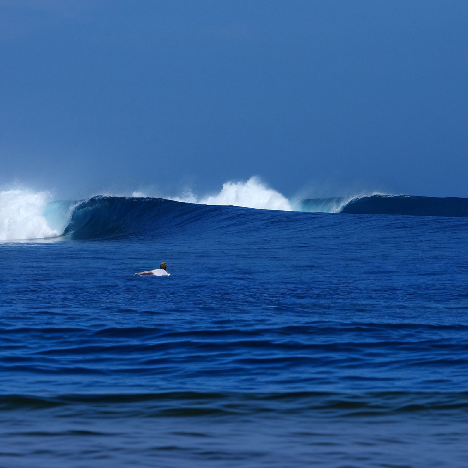 Mar azul profundo com séries de ondas quebrando ao fundo e um surfista distante remando